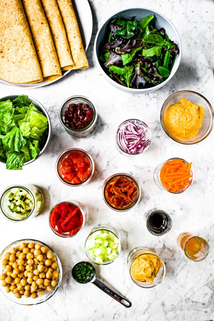 Overhead shot of the ingredients you need to make a mediterranean hummus wrap: sun-dried tomato tortillas, hummus, mixed greens, romaine, roasted red peppers, sun-dried tomatoes, kalamata olives, chickpeas, red onions, cucumbers, pickles, balsamic vinegar, shredded carrots, minced parsley