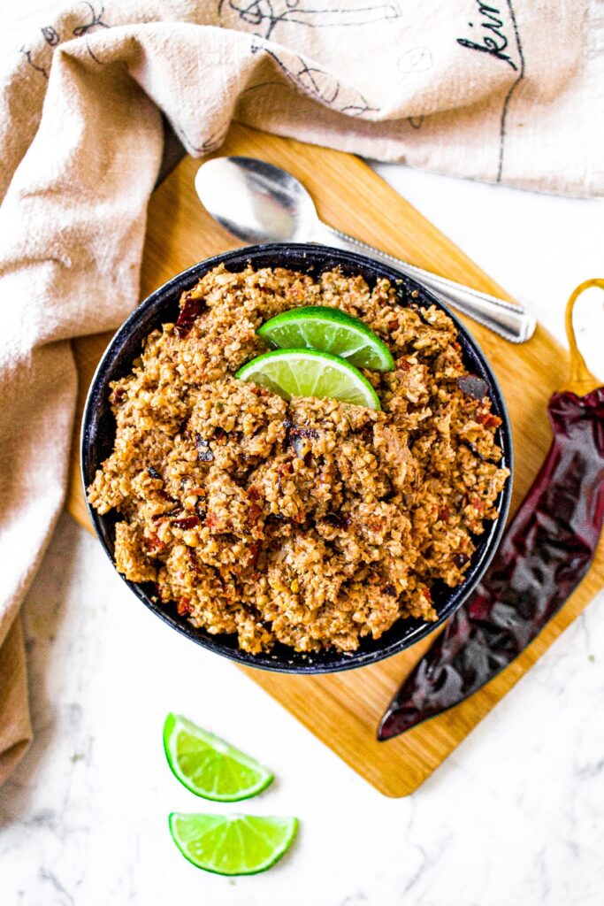 Overhead shot of a bowl of chorizo style walnut meat topped with 2 lime wedges. There are more lime wedges around the bowl and a long guajillo chili pepper is next to the bowl.