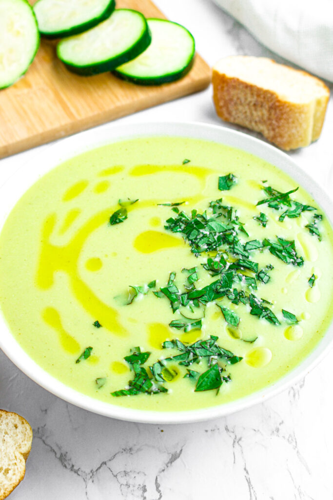 Overhead view of a round white bowl of creamy vegan zucchini soup topped with minced herbs on the right side