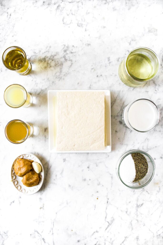 Overhead shot of all the ingredients you need to make vegan goat cheese: extra firm tofu block, miso paste, apple cider vinegar, lemon juice, water, non-dairy milk, salt, pepper, and refined coconut oil