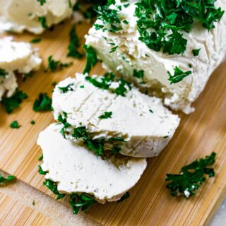 Close up overhead shot of a tube of vegan goat cheese with 2 slices sliced. There is another log of dairy free goat cheese next to it and minced parsley on top.