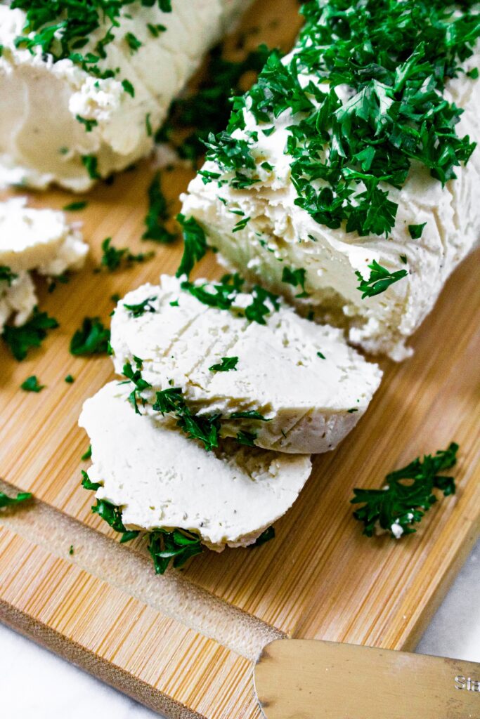 Close up overhead shot of a tube of vegan goat cheese with 2 slices sliced. There is another log of dairy free goat cheese next to it and minced parsley on top.