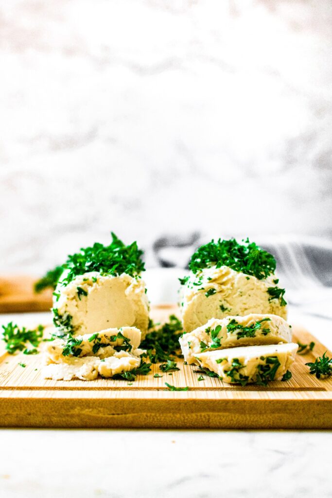 Head on shot of two tubes of easy vegan goat cheese that has been sliced twice. The slices lie flat in front of the intact tubes. Each is sprinkled with fresh parsley and sitting on a wooden cutting board